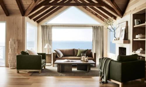 Bright living room with wooden beams, green armchairs, and ocean views through large windows.