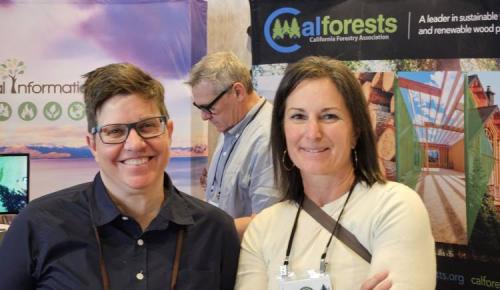 Two conference attendees smile together in front of a Calforests exhibit booth showcasing sustainable and renewable wood products at the California Forestry Association meeting.