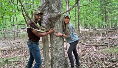 Two people smile and wrap their arms around a large tree trunk in a leafy forest.