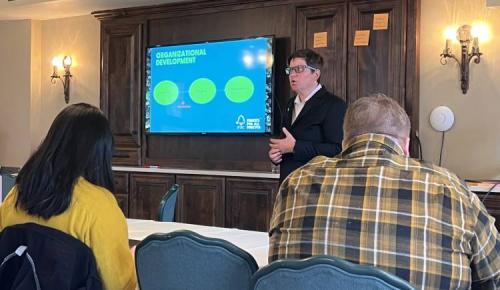 A presenter speaks beside a screen displaying a slide titled “Organizational Development” as participants listen.