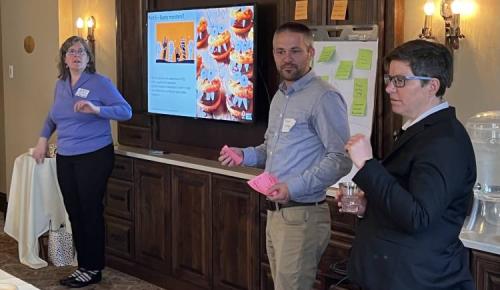 Three presenters stand near a screen and flip chart while engaging participants during a discussion.