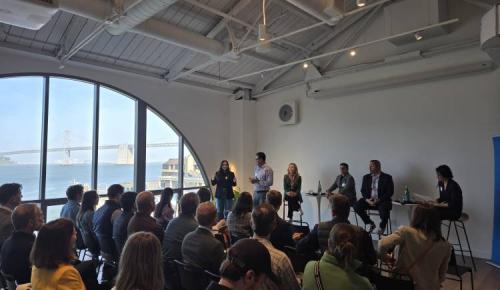 A large group listens to presenters at a waterfront venue with tall arched windows overlooking the San Francisco–Oakland Bay Bridge.