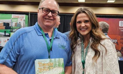 Two people smile at an event, one holding a children’s book titled Travis Visits the Tree Farm.