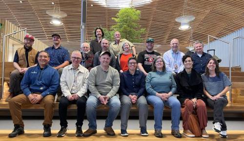 A large group sits and stands on wooden steps inside a modern building with a curved wood ceiling.