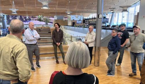 Participants stand in a circle listening to a speaker inside a large, open building with wooden floors and ceiling.