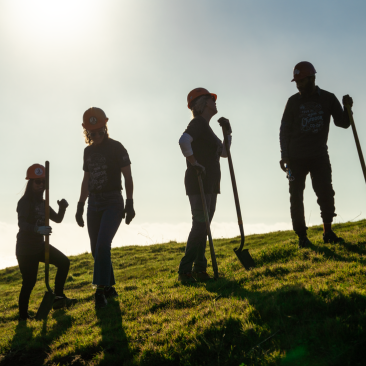 Four volunteers wearing outdoor gear and hardhats standing on a grassy knoll with shovels at sunset.